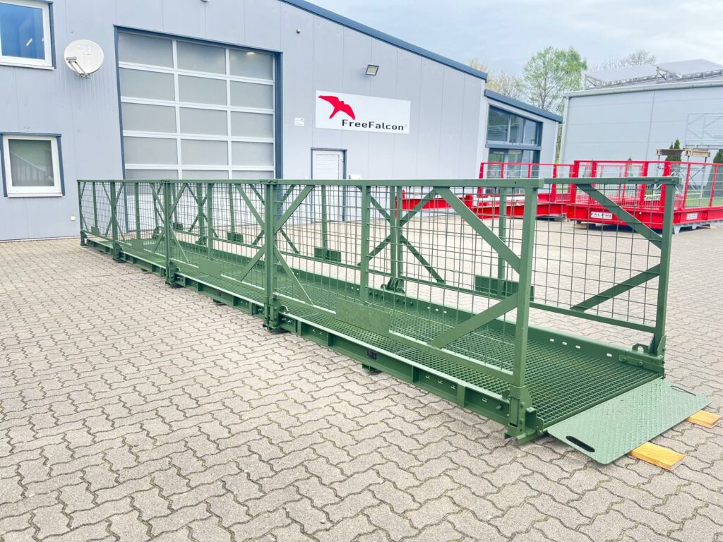 Eine tragbare Fußgängerbrücke aus Metall mit grünem Geländer steht auf einer gepflasterten Fläche vor einem Industriegebäude mit einem „FreeFalcon“-Schild.
