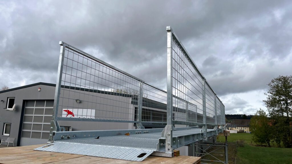 An elevated walkway made of metal mesh and rails over a wooden platform in an industrial area, with overcast sky and trees in the background.