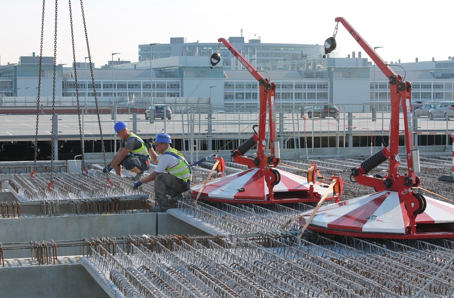 Construction workers install steel reinforcements at a building site, using cranes and heavy machinery, while undergoing training in proper techniques, with an industrial facility in the background.