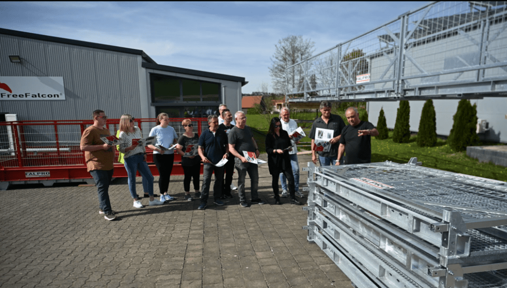 A group of people stands outside a building, facing metal equipment. Some hold documents while one person gestures towards the equipment, indicating a training session in progress. The scene appears to be a professional or instructional setting.