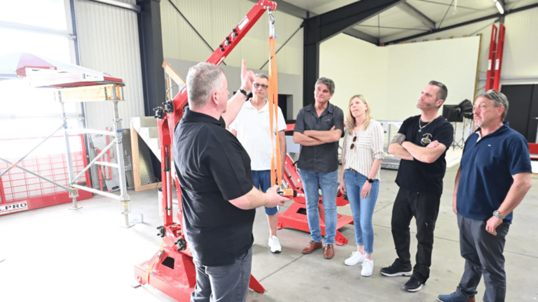 A group of six people stands in a workshop, attentively listening to a person explaining something beside a red mechanical apparatus. The "About Us" section highlights the collaborative spirit and expertise found in our well-lit, industrially equipped workspace.