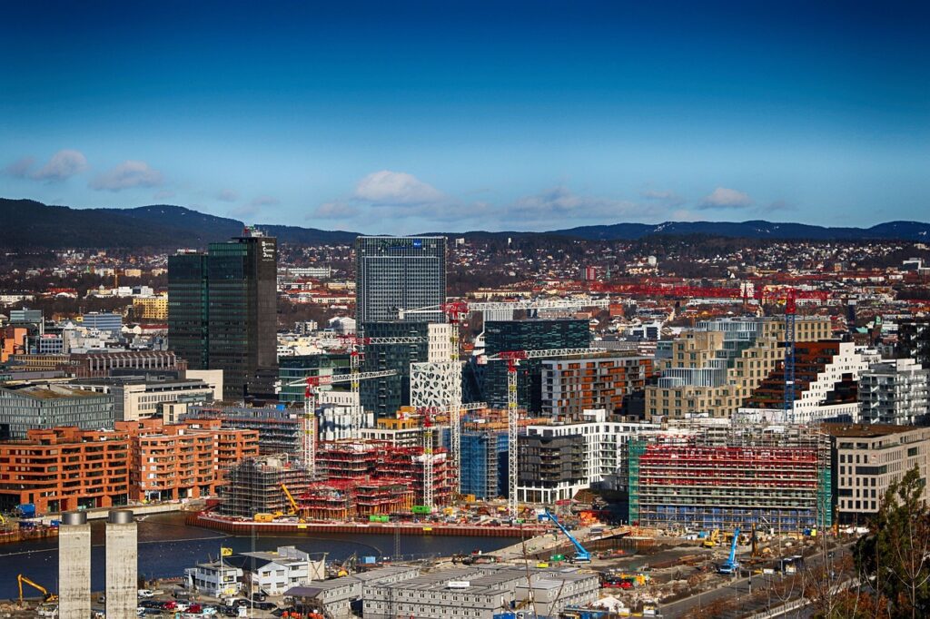 Eine Stadtansicht eines modernen Stadtgebiets mit mehreren Hochhäusern, Baustellen und einem Fluss im Vordergrund, vor einer Hügelkulisse unter einem teilweise bewölkten Himmel, spiegelt das dynamische Wachstum wider, das in Metropolen weltweit zu beobachten ist.