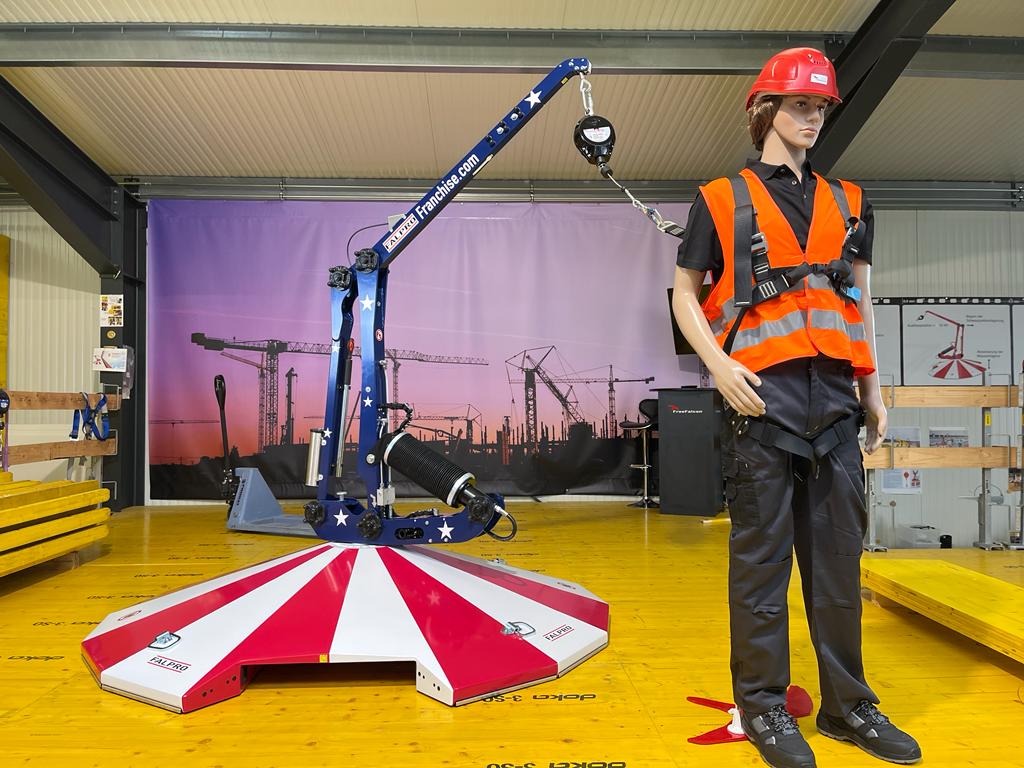 A mannequin wearing a red safety helmet, orange vest, and harness stands next to a blue portable crane with a red-and-white base in an indoor training facility.