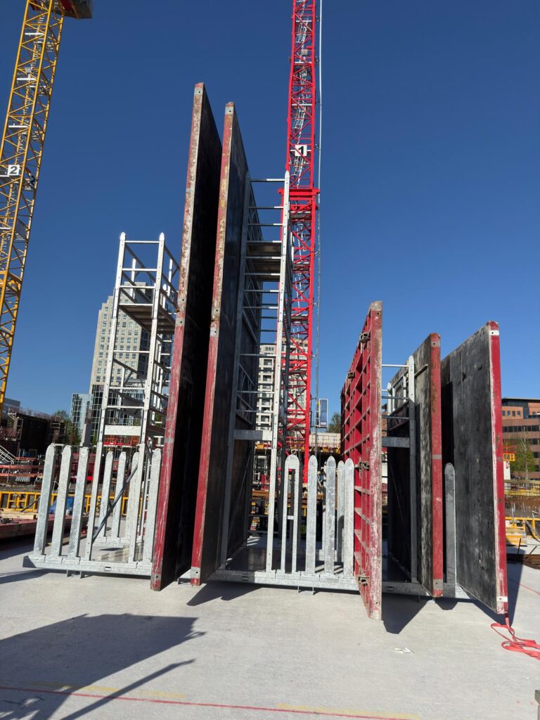 Several large concrete formwork panels stand upright on a construction site, organized with a panel storage rack, while cranes and buildings are visible in the background under a clear blue sky.